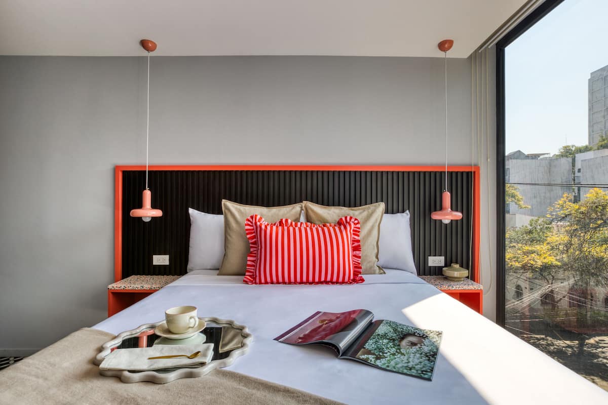 A modern hotel room with a large bed, two bedside tables, a window with a view of the city, and a tray with a cup and book on the bed.