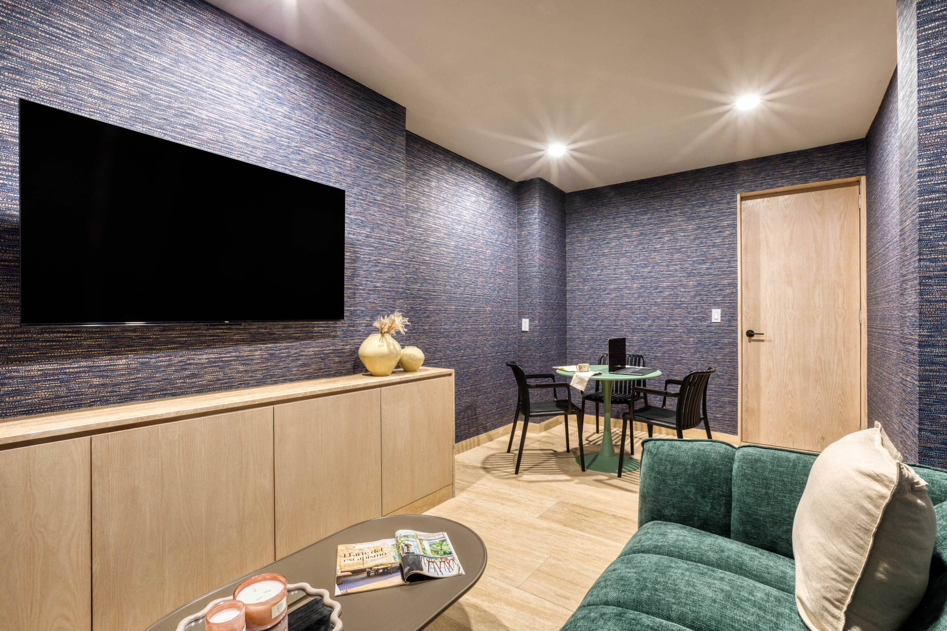 Una sala de estar moderna con un televisor de pantalla plana montado en la pared, un sofá verde, una mesa de centro con revistas y un comedor con una mesa pequeña y sillas.