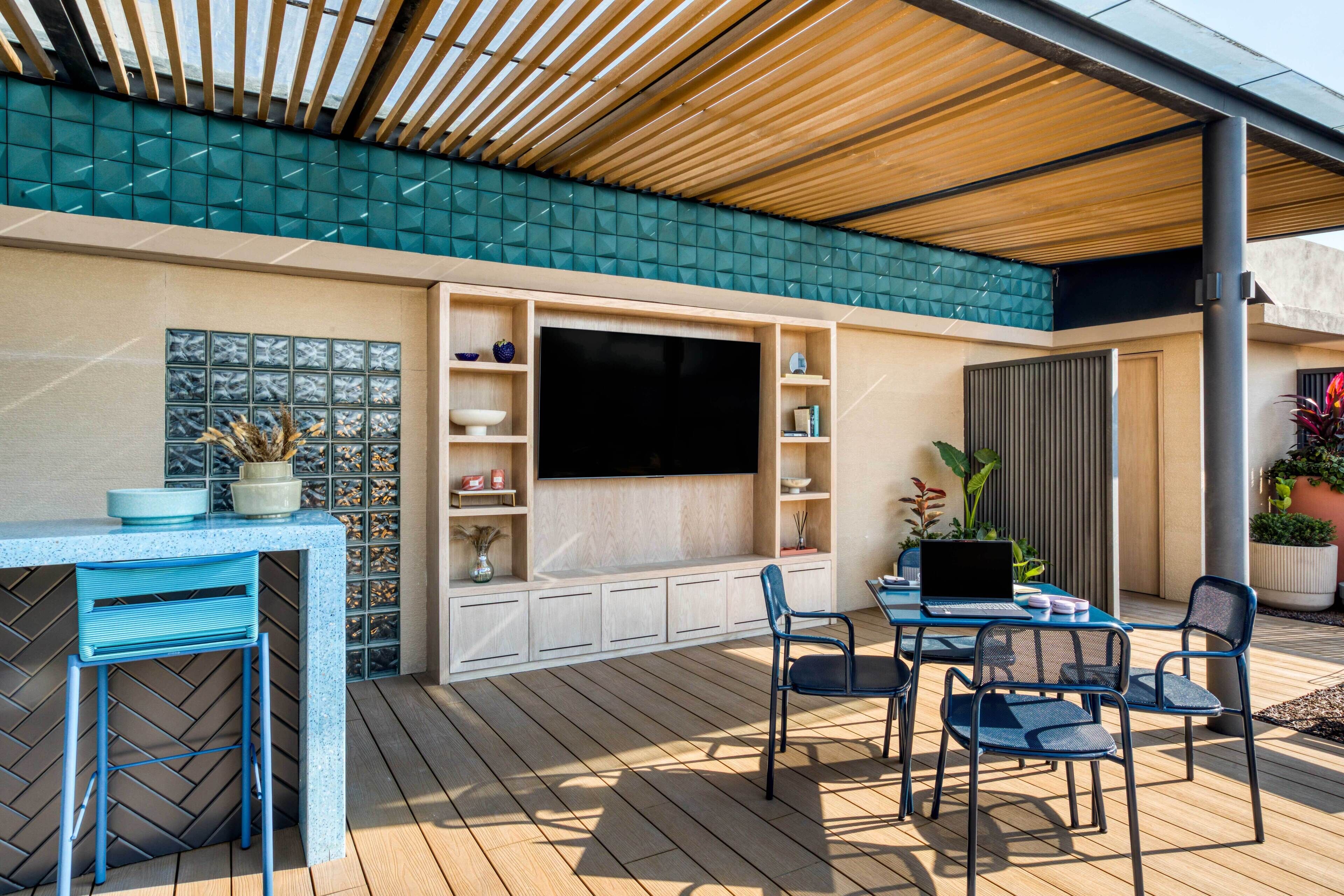 Un patio al aire libre con techo de madera, un televisor montado en un mueble blanco, y una mesa con sillas.