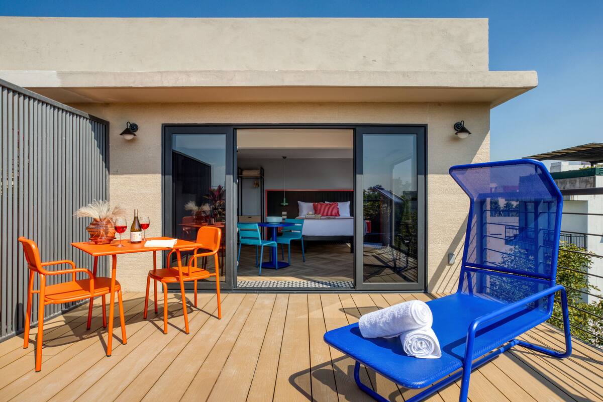 An outdoor deck with a table and chairs, a blue lounge chair, and a sliding glass door leading to a bedroom with a bed and a desk.