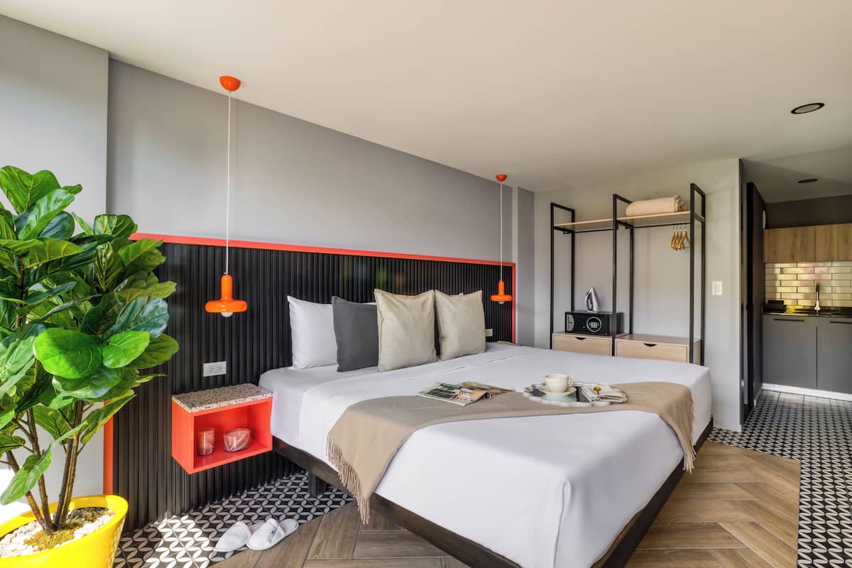 A modern hotel room with a large bed, a small desk, a potted plant, and a kitchen area visible through an open door.