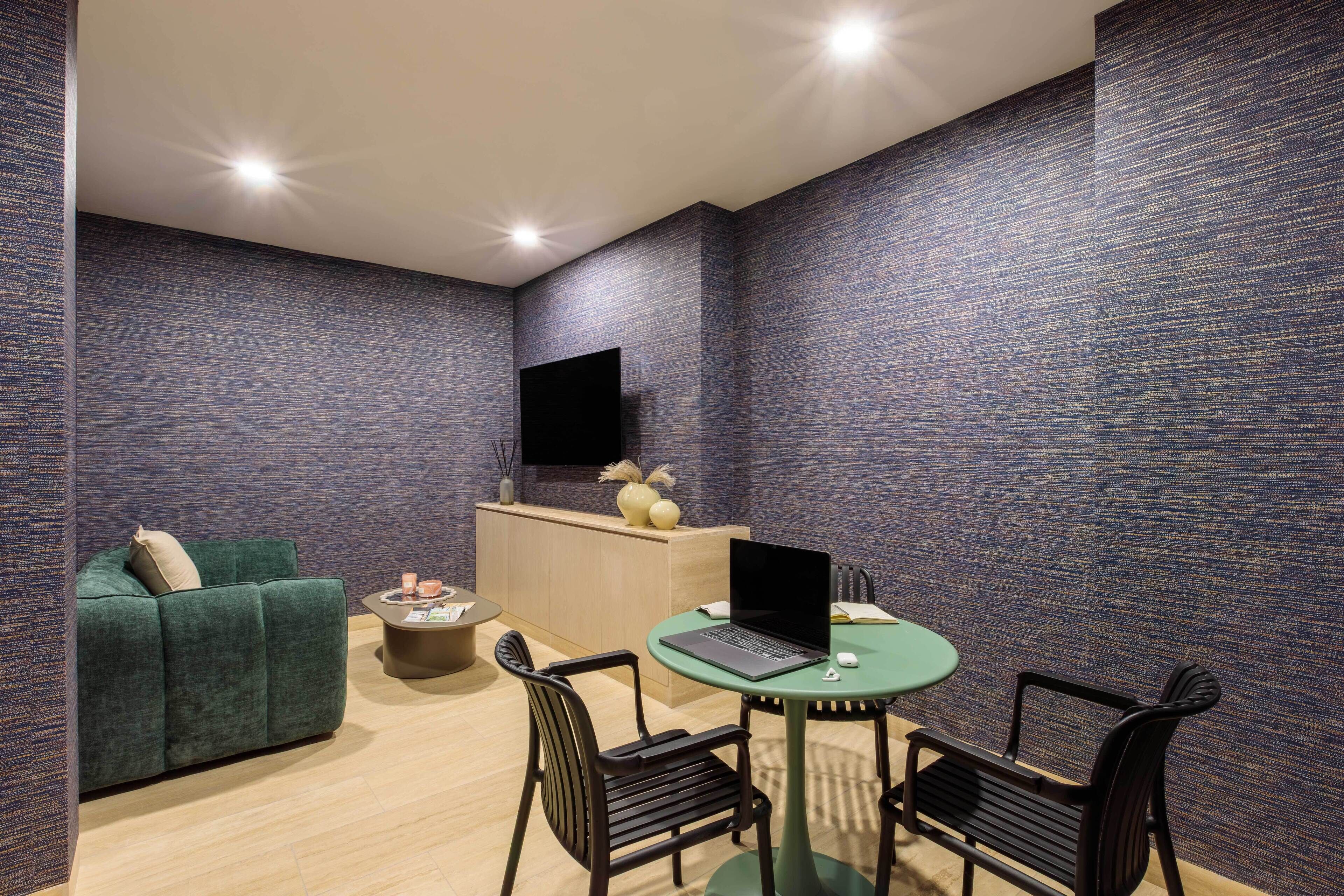 A modern room with a green sofa, a small round table with a laptop, two black chairs, a wooden cabinet, and a flat-screen TV mounted on the wall.
