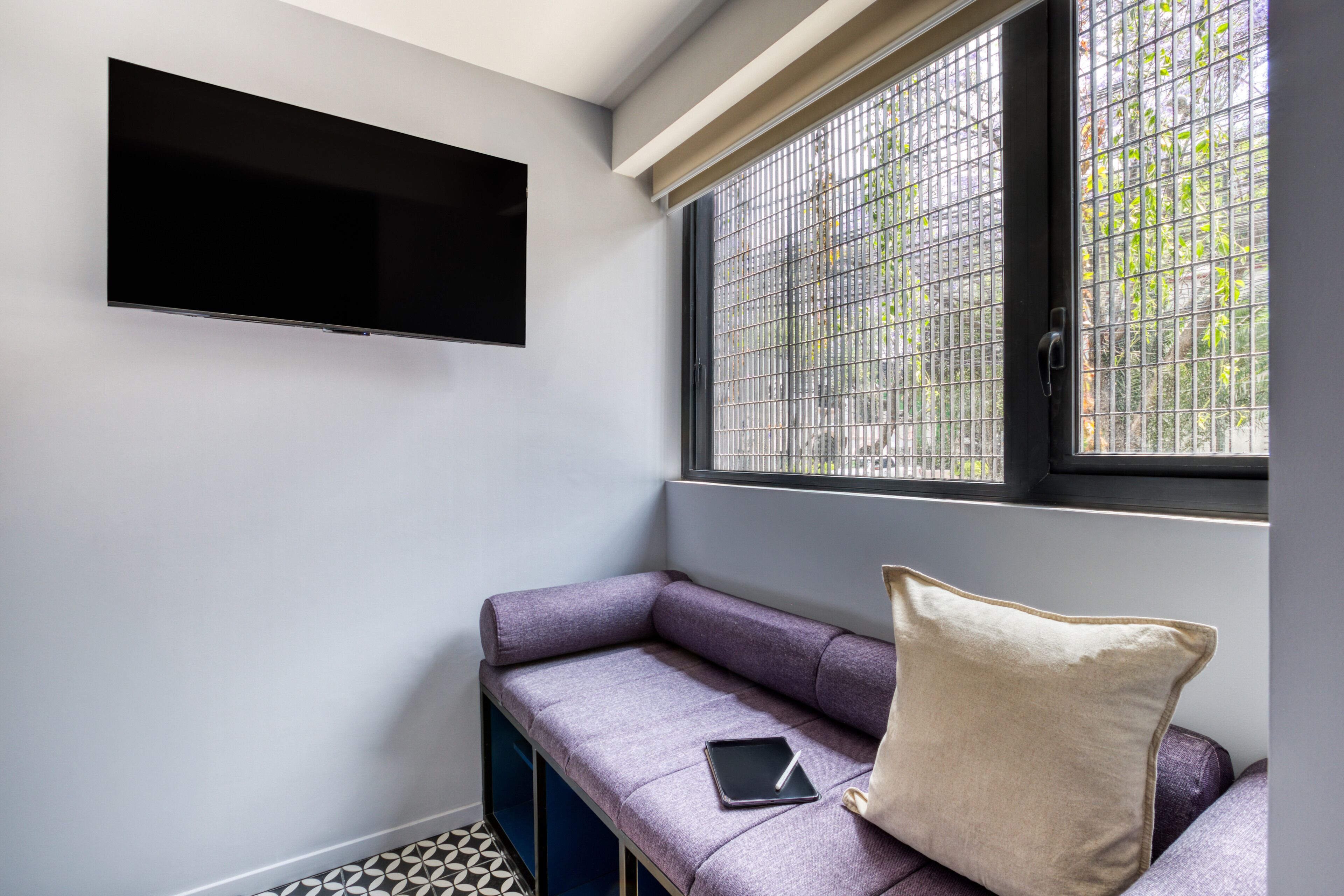 Una habitación moderna con televisor integrado, ventana con rejas de metal, sofá púrpura con una tableta y un almohadón, y piso con estampado.