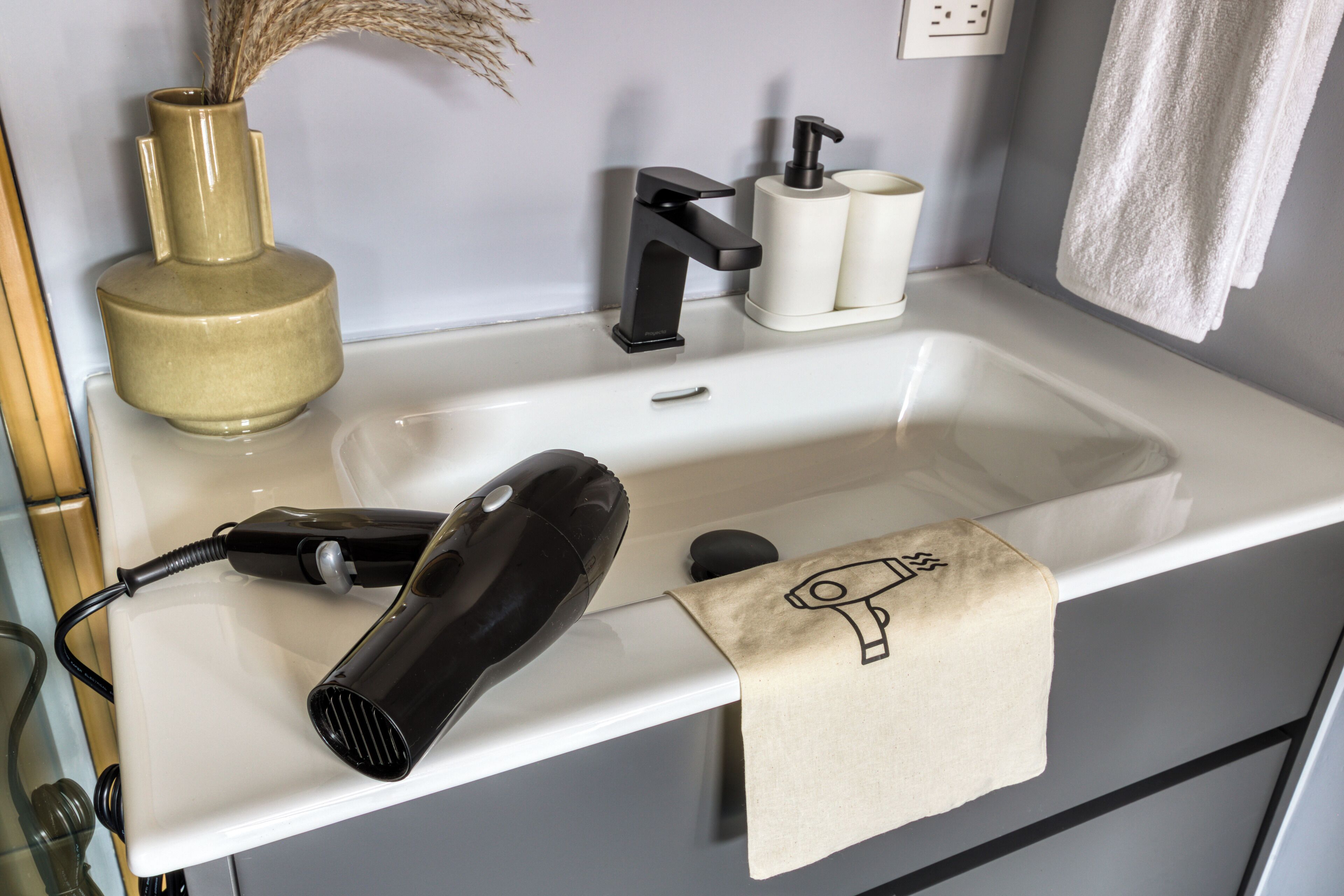 A bathroom with a white sink, a black hairdryer, a beige towel, and a vase with dried plants.