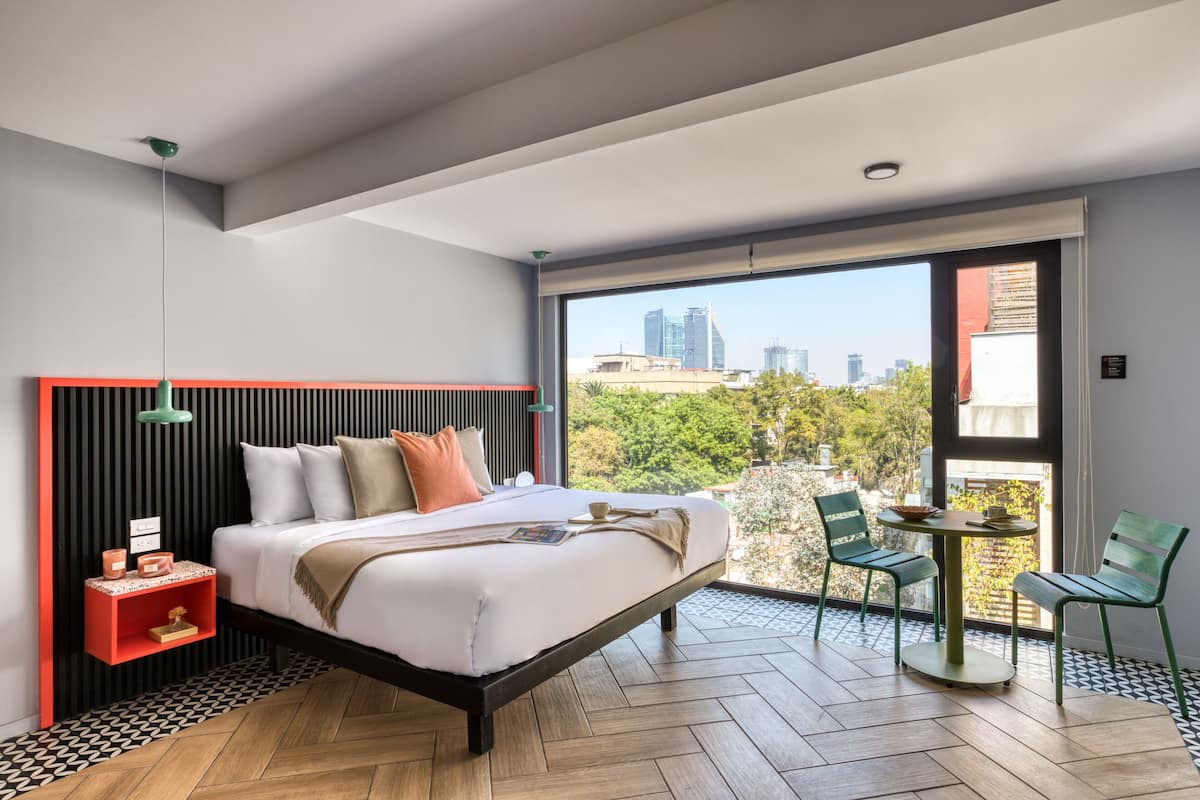 A modern bedroom with a city view, a wooden floor, a bed with pillows, a nightstand, and a small table with chairs.