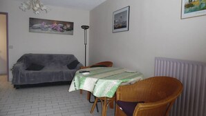 A room with a grey sofa, a dining table with a green tablecloth, wicker chairs, a floor lamp, and a radiator. - Bright 2-room apartment with private parking at Canet-Plage (Canet-Plage)