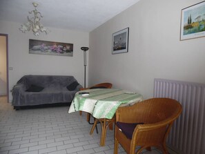 Ein Zimmer mit einem grauen Sofa, einem Esstisch mit grüner Tischdecke, Korbstühlen, einer Stehlampe und einem Heizkörper.