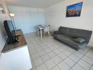 A compact living space with a grey sofa, a dining table with chairs, a TV on a stand, and a painting on the wall. - Renovated 2-bedroom apt facing sea, balcony, 4 pers (Canet-Plage)