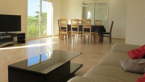 Una sala de estar con televisor, un sofá, una mesa de comedor con sillas y un ventanal corredizo que da a un balcón.