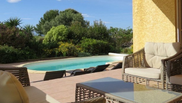 Zona junto a la piscina con mobiliario de mimbre, una mesa de vidrio y vista a una alberca rodeada de abundante vegetación.