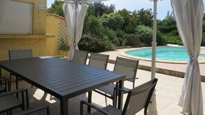 Un patio exterior con mesa y sillas, un área techada con piscina y vegetación circundante.