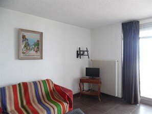 A living room with a striped sofa, a television on a wooden stand, a radiator, and a window with curtains. - Air-conditioned two-room sea view, near port (Canet-Plage)