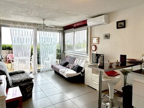 Un espace de vie compact comprenant un canapé, une table à manger, un coin cuisine et une porte-fenêtre coulissante donnant sur un espace extérieur.