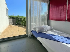A single bed with white bedding and a blue blanket, a window with sheer curtains, and a view of a garden and a building outside. - Beachfront Studio with A/C, Parking & Terrace (Le Barcarès)