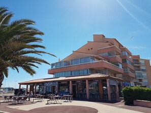 A multi-story building with a bar and restaurant on the ground floor, outdoor seating, and a palm tree in the foreground. - Pleasant studio with terrace, beach access, private parking (Le Barcarès)