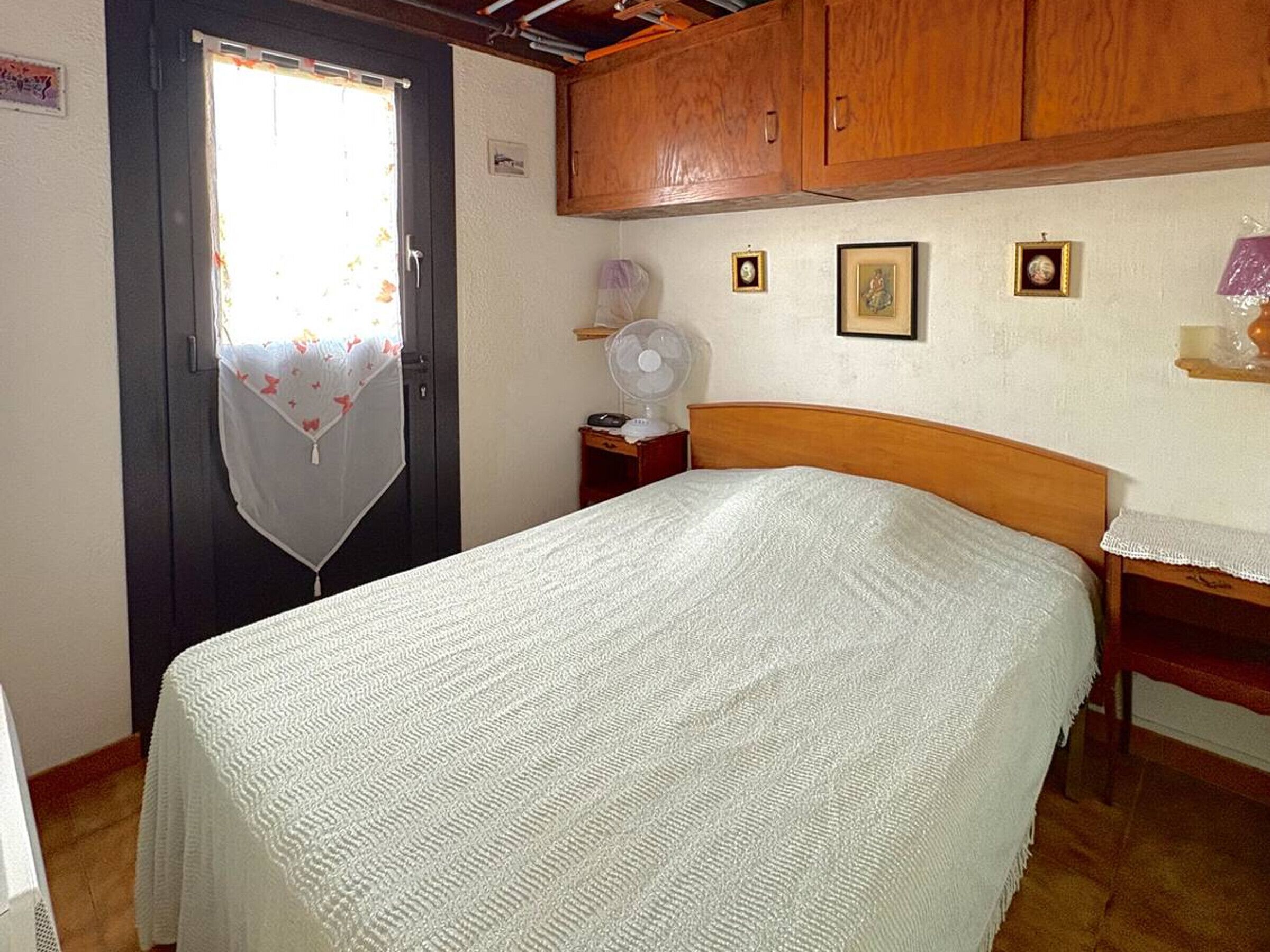 A bedroom with a bed, wooden wardrobe, a small bedside table, a fan, and a door with a curtain.