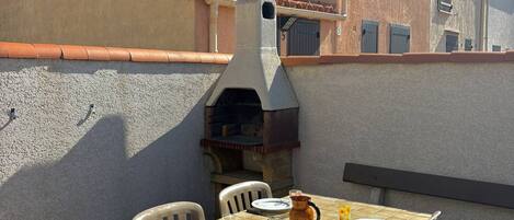 An outdoor dining area with a stone table and chairs, a stone oven, and a building with a tiled roof in the background.
