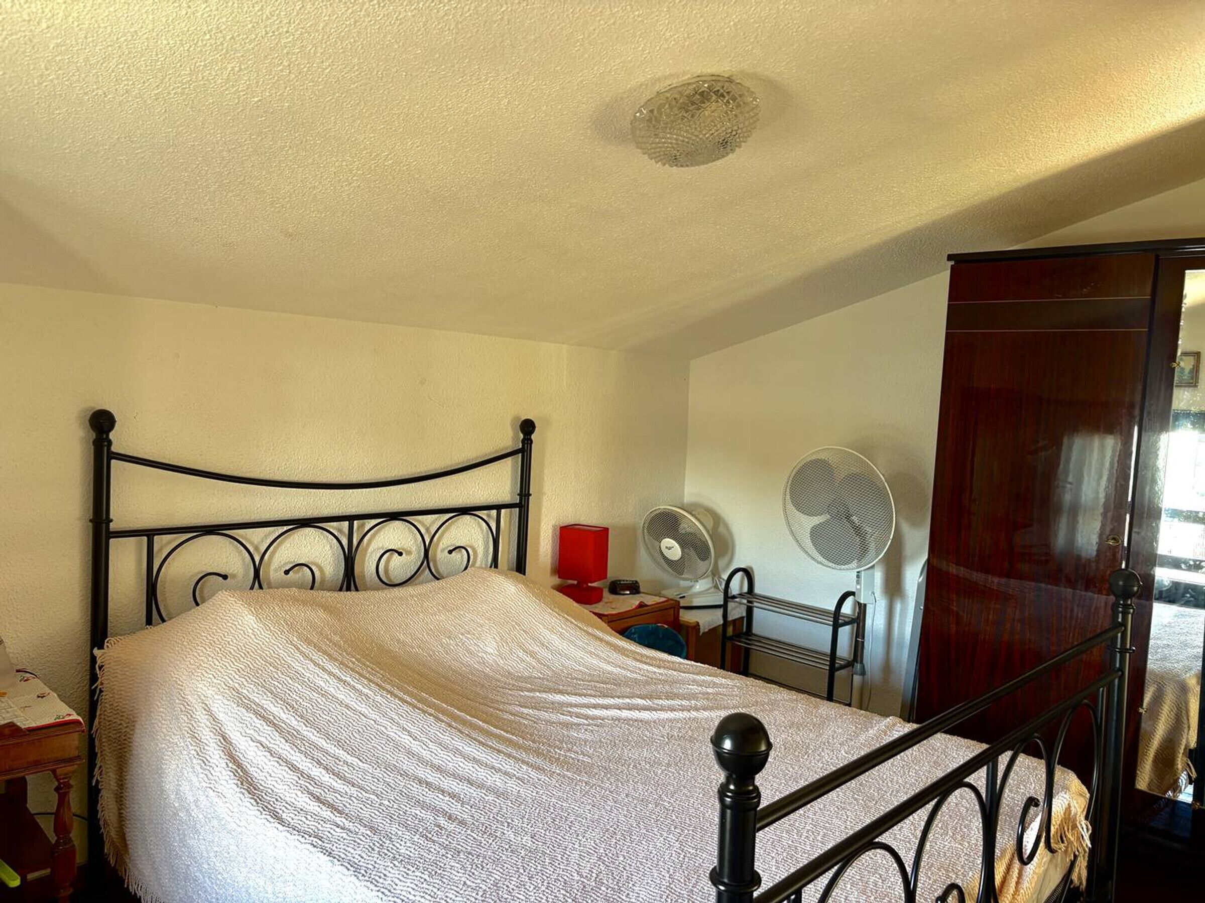 Uma cama de solteiro com estrutura de metal em um quarto com paredes brancas, uma porta de madeira e duas ventoinhas.