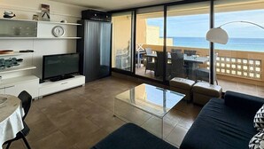 A modern living room with a flat-screen TV, a glass coffee table, and a sofa. There is a balcony with a view of the sea. - Seaside apartment with terrace and secure parking (Le Barcarès)