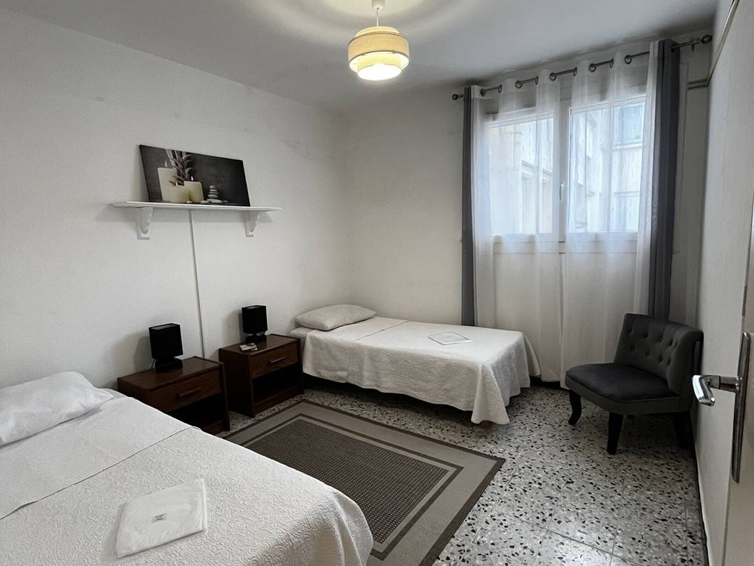 A hotel room with two beds, a chair, a desk with a lamp, a shelf with a picture, and a window with curtains.