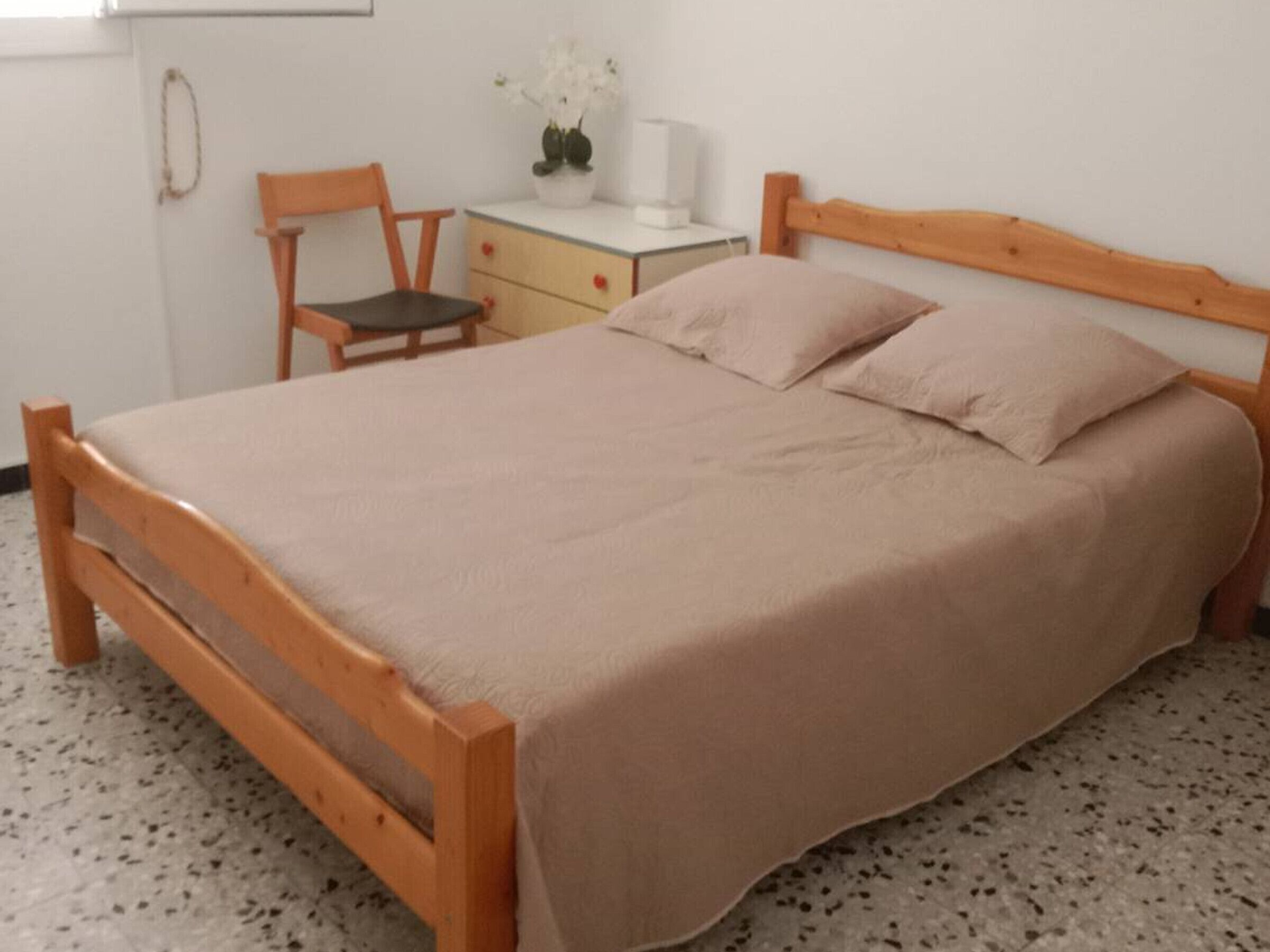 A bedroom with a wooden bed, a chair, a small table with a vase of flowers, and a nightstand.
