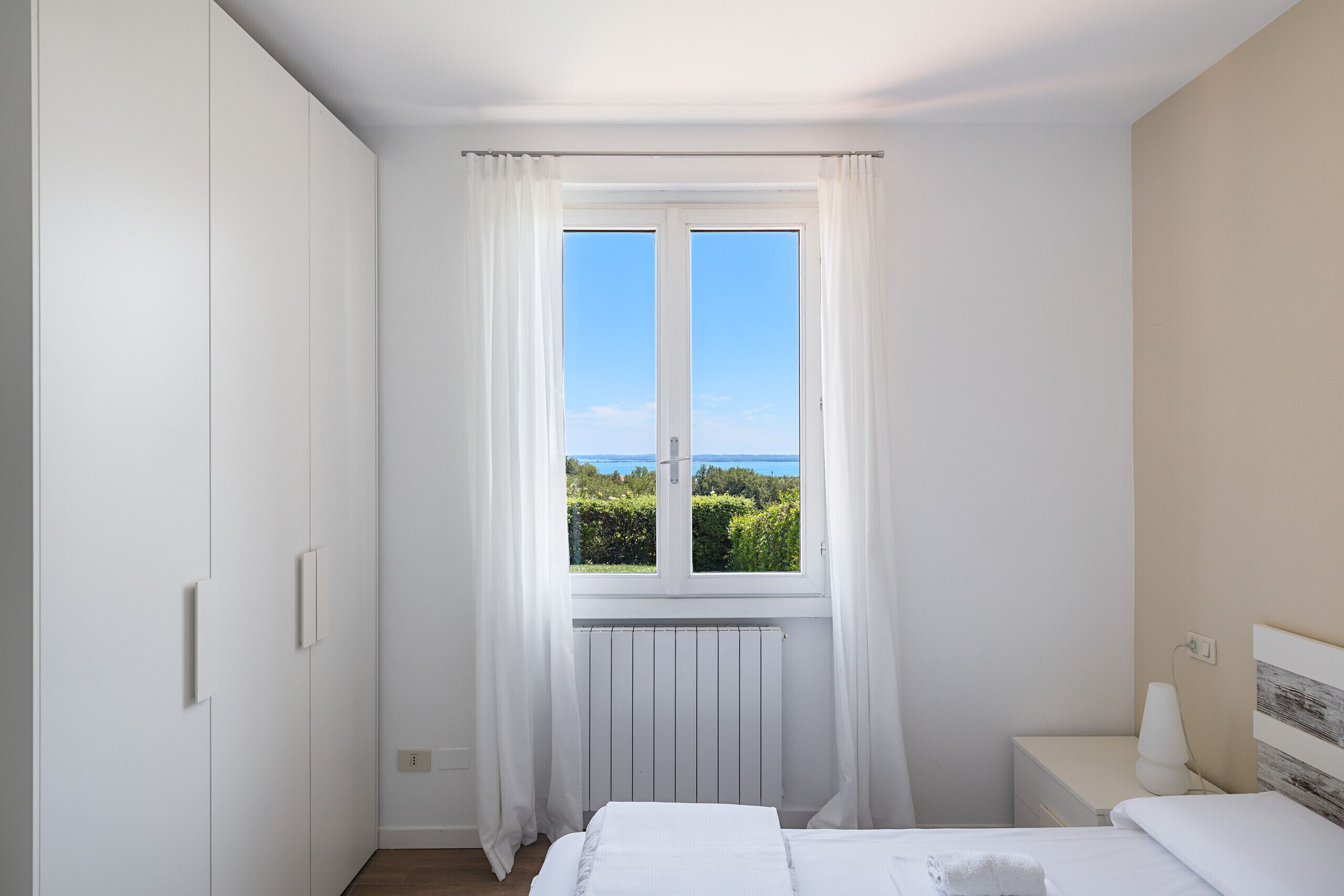 Un dormitorio con cama, mesita de noche, ventana con vistas a la vegetación y al agua, y un radiador.