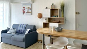 Ein modernes Wohnzimmer mit einem grauen Sofa, einem Holzesstisch und weißen Stühlen. An der Wand hängt ein Bild, eine Zimmerpflanze steht bereit und eine Tür ist mit einem Schloss gesichert.