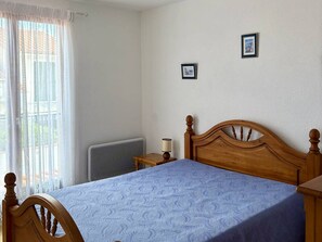 A bedroom with a wooden bed, a radiator, a bedside table with a lamp, and a window with sheer curtains. - Cozy House with Pool, 500m from Beach, Sleeps 5, Le Barcarès (Le Barcarès)