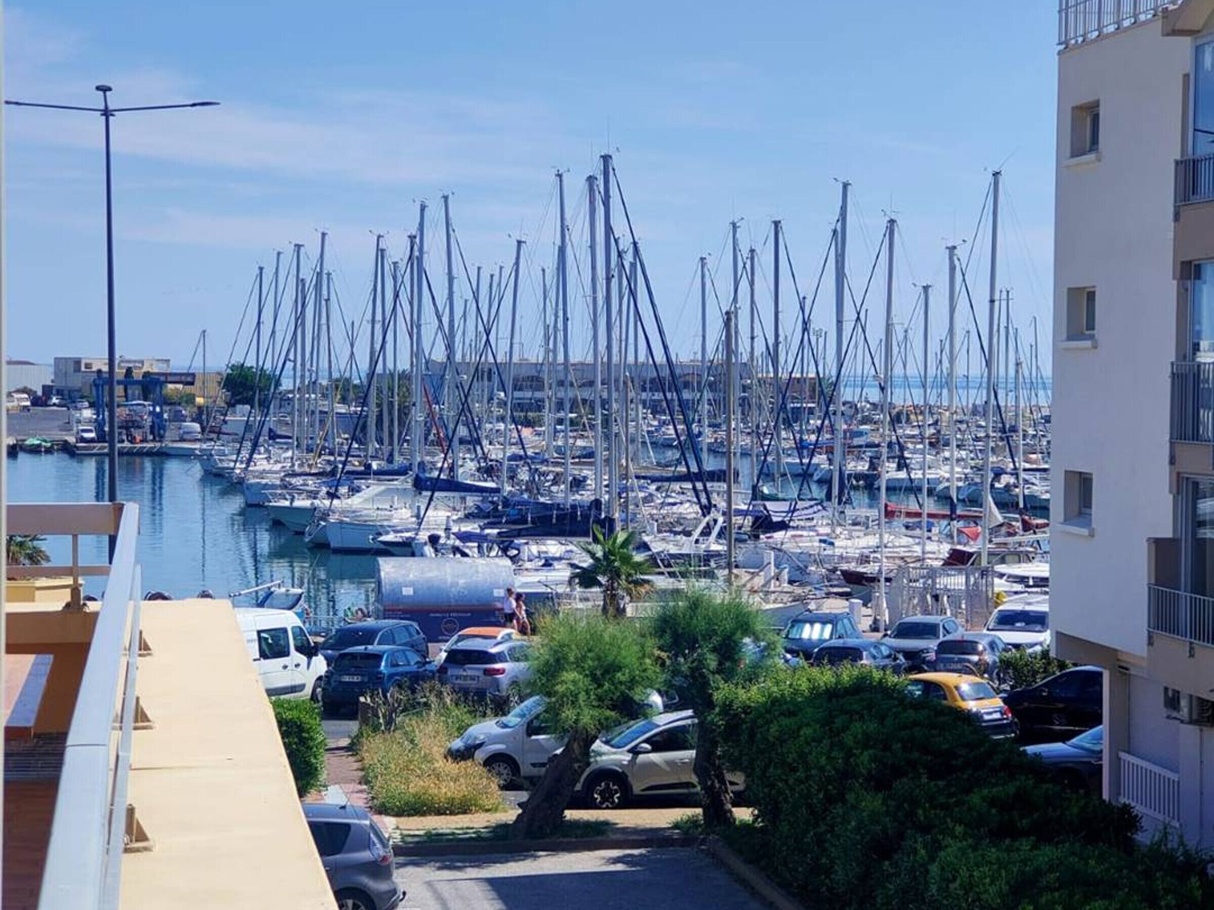 Un port de plaisance où sont amarrés de nombreux bateaux, un parking avec des voitures et un immeuble doté de balcons.