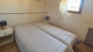 Una habitación pequeña con cama individual, mesita de noche con lámpara, ventilador de pared y ventana con cortinas.