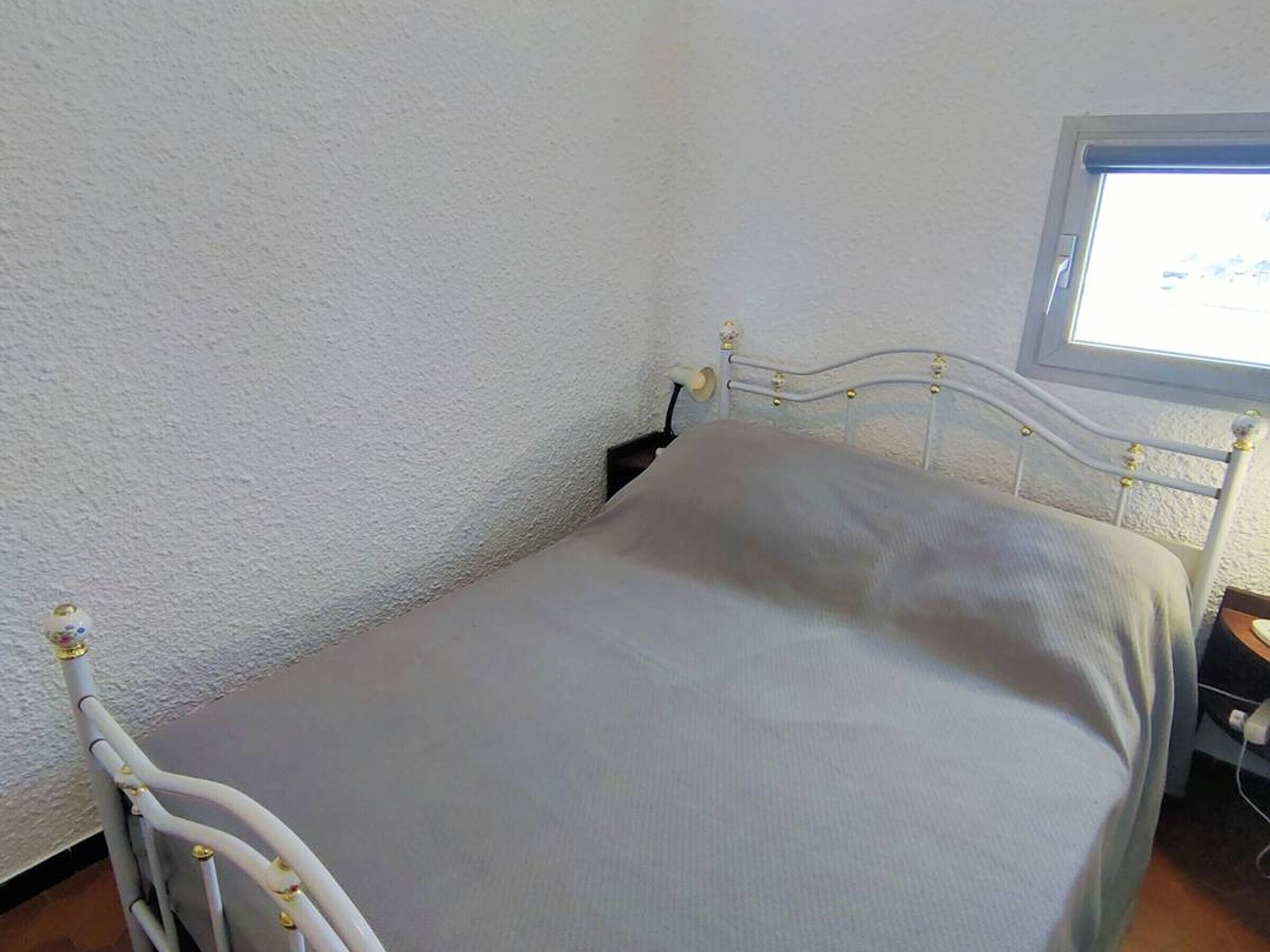 A single bed with a white metal frame and a grey mattress in a room with white walls and a window.