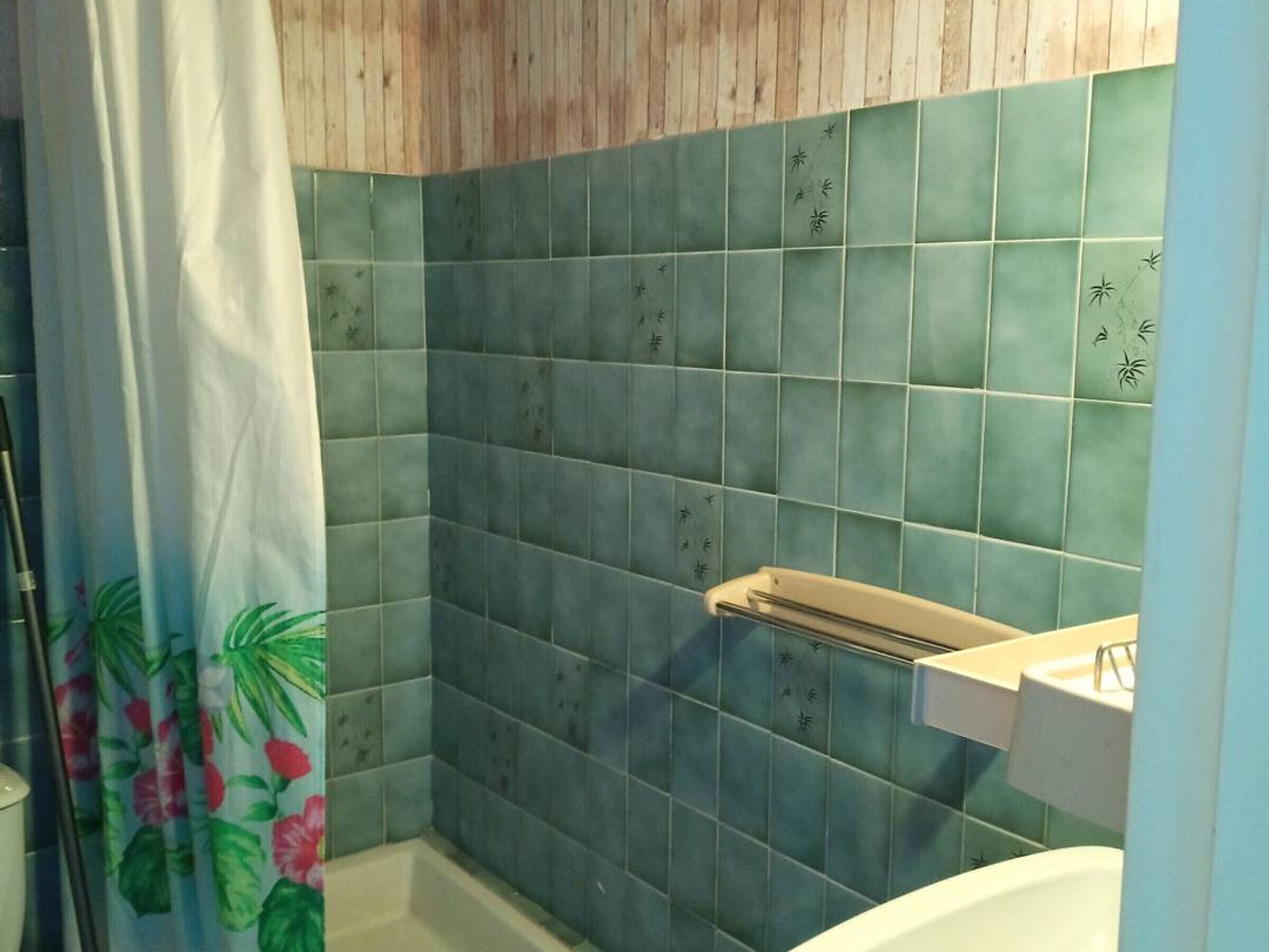 Un baño con azulejos verdes, una cortina de ducha con motivos florales y una bañera blanca.