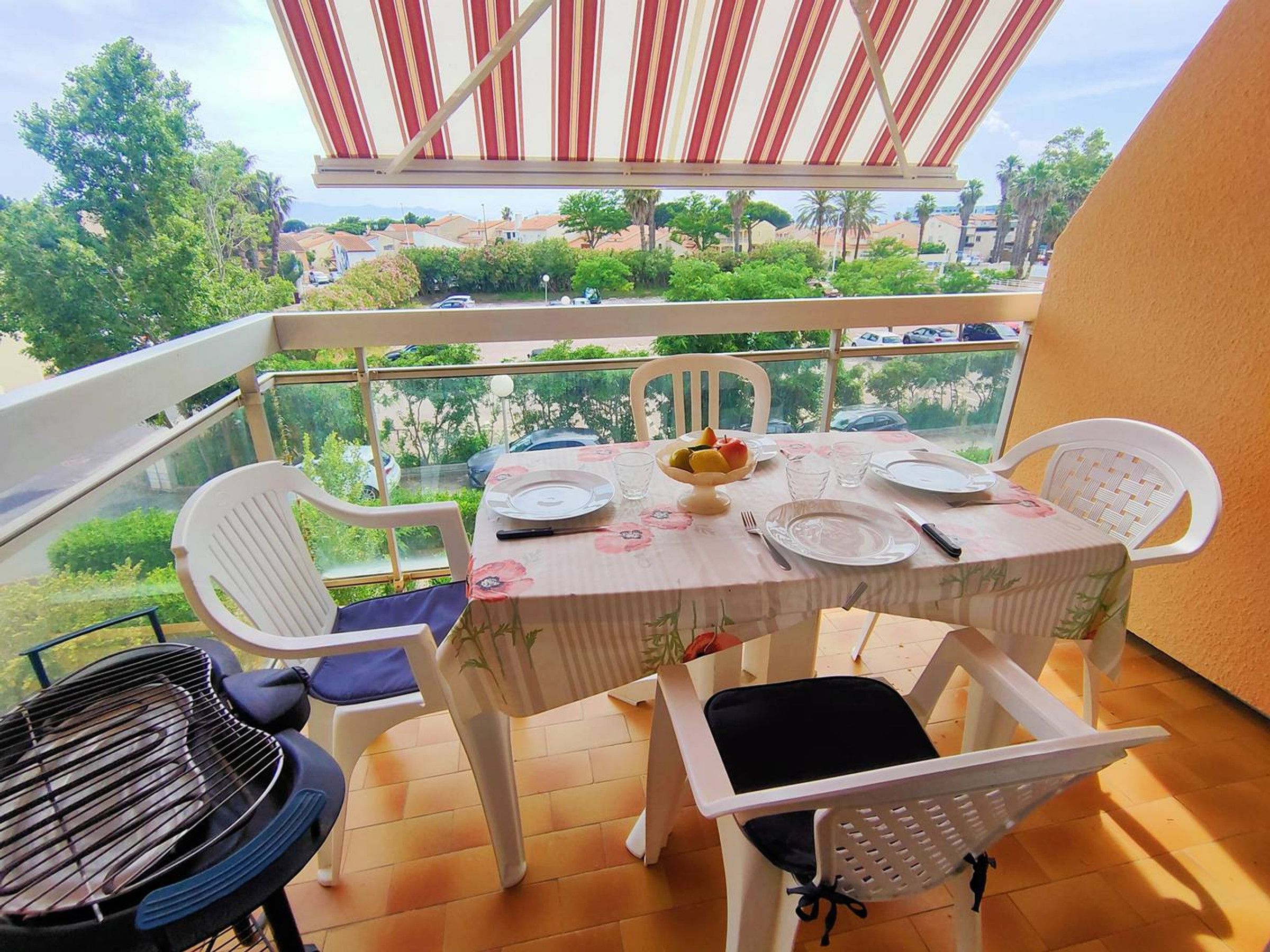 A balcony with a table set for two, chairs, a grill, and a view of a residential area.