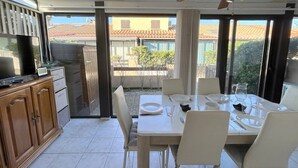 Ein Essbereich mit Tisch für vier Personen, eine Küche mit Schränken und einem Fernseher, sowie eine Glastür, die auf eine Terrasse mit Blick auf ein Haus und Bäume führt.