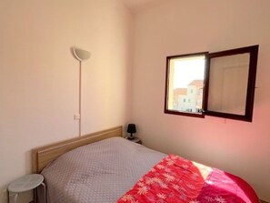 Ein Schlafzimmer mit Bett, Nachttisch, Lampe, Fenster mit Blick auf Gebäude und Wandleuchte.