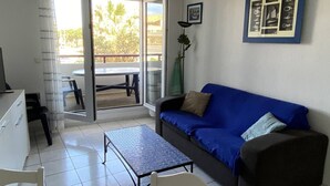 Sala de estar com um sofá azul, uma mesinha e uma cadeira. Há uma porta de vidro deslizante que dá para uma área externa com uma mesa e cadeiras.
