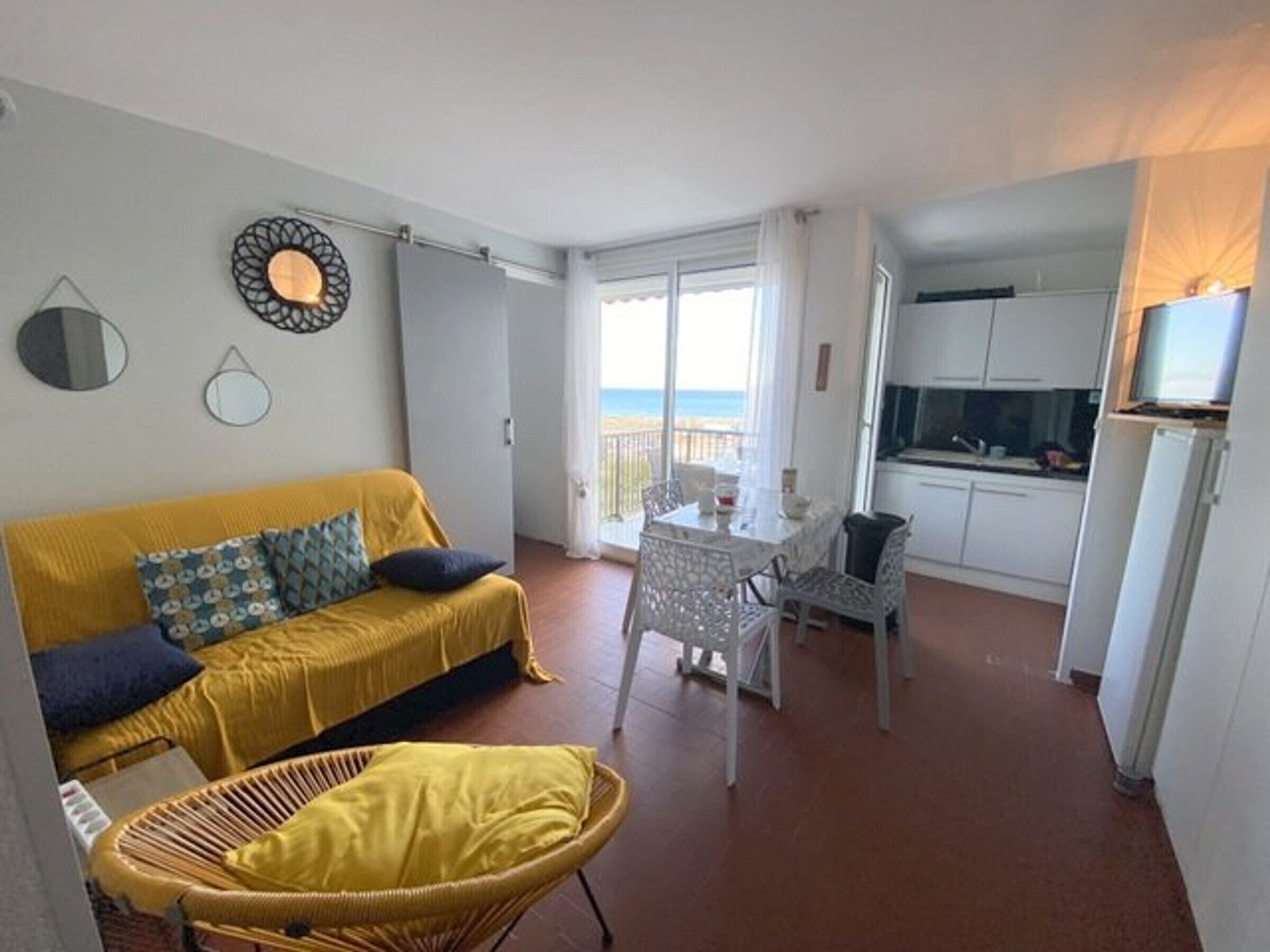 A compact living space with a yellow sofa, a wicker chair, a dining table, and a kitchen area.