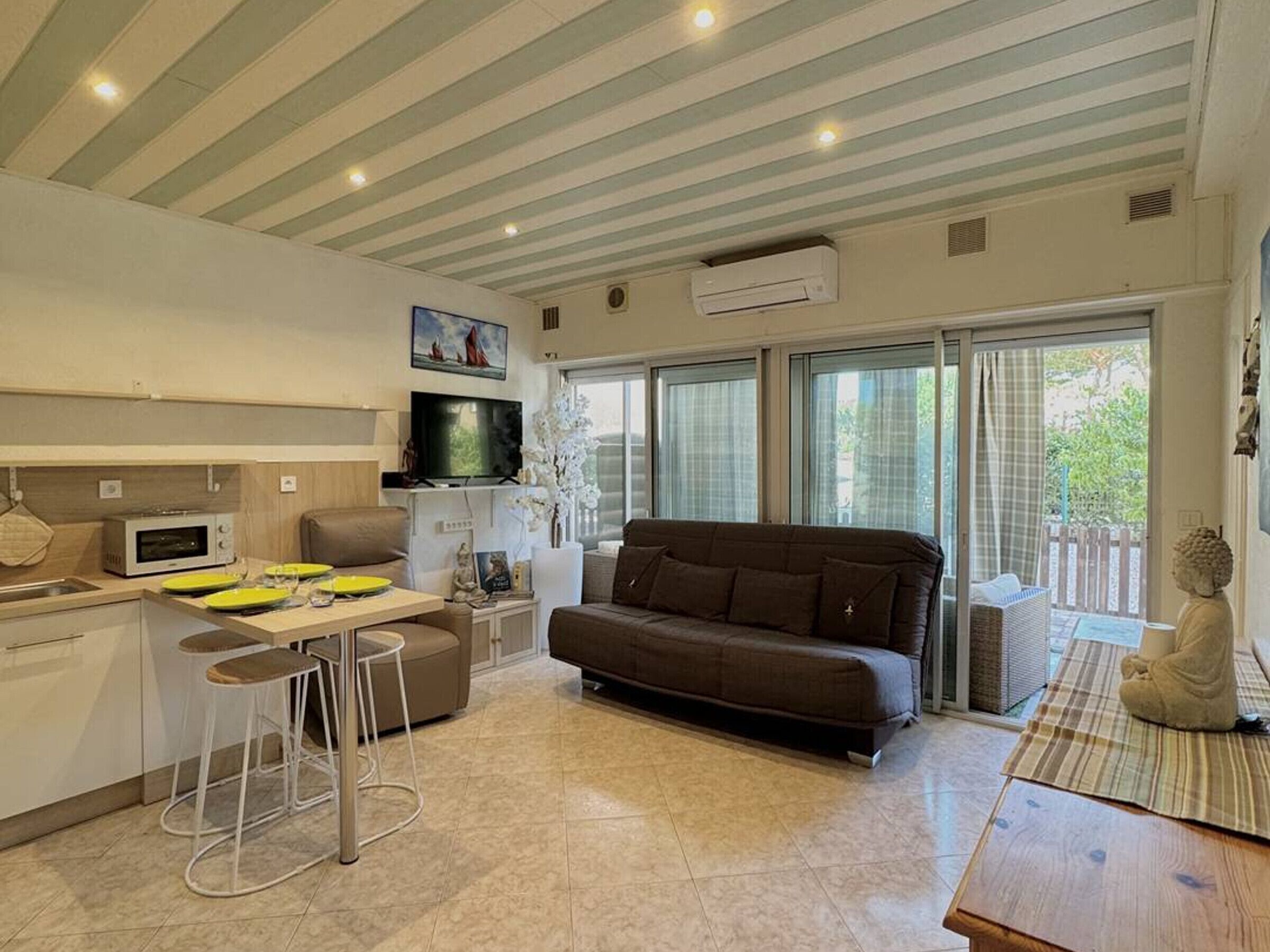 A modern kitchen with a dining area, a sofa, and a large window with a view of greenery.