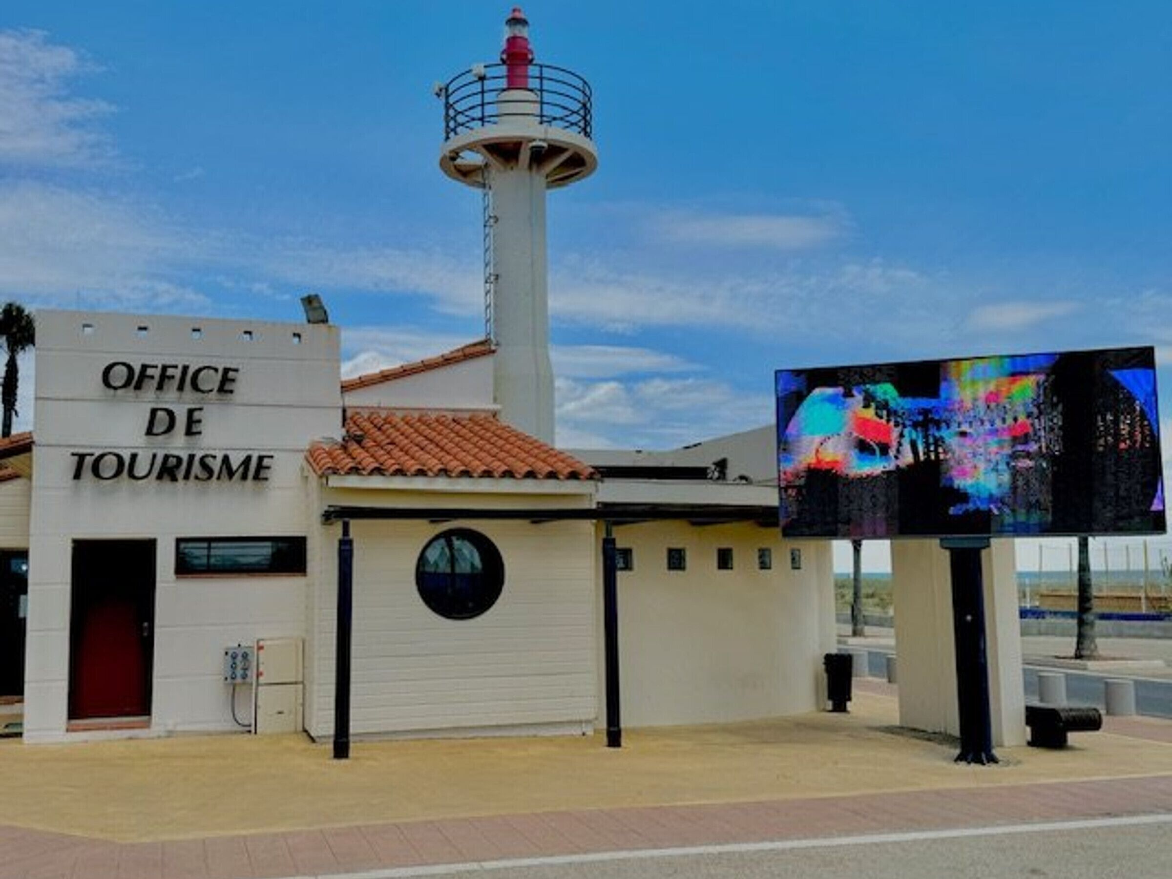 A building labeled "OFFICE DE TOURISME" with a lighthouse-like structure, a large digital billboard, and a paved walkway.