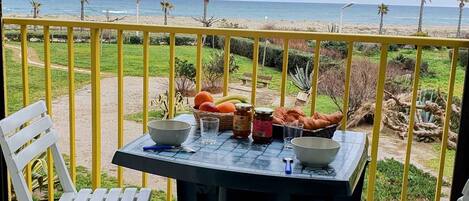 A balcony with a table set for two, overlooking a beach with palm trees and the ocean.
