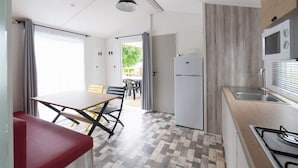 A compact kitchen with a gas stove, a built-in microwave, and a refrigerator. There is a dining area with a table and chairs, and a view of the outdoors through a door. - Cozy mobile home with terrace for 4 in Bénodet (Bénodet)