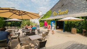 An outdoor patio area with tables and chairs, a wooden deck, and a building with a sign reading "Car Saek". - Cozy mobile home with terrace for 4 in Bénodet (Bénodet)