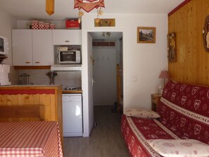 Eine kompakte Küche mit Mikrowelle, Toaster und kleinem Kühlschrank. Im Vordergrund steht ein Esstisch mit kariertem Tischläufer. Im Hintergrund ist eine rote Couch mit Herzmustern zu sehen.