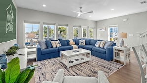 A modern living room with a blue sectional sofa, a coffee table, and large windows offering a view of the outdoors. - Almost Heaven on Sunset-Pool-Walk to Beach (Murrells Inlet)