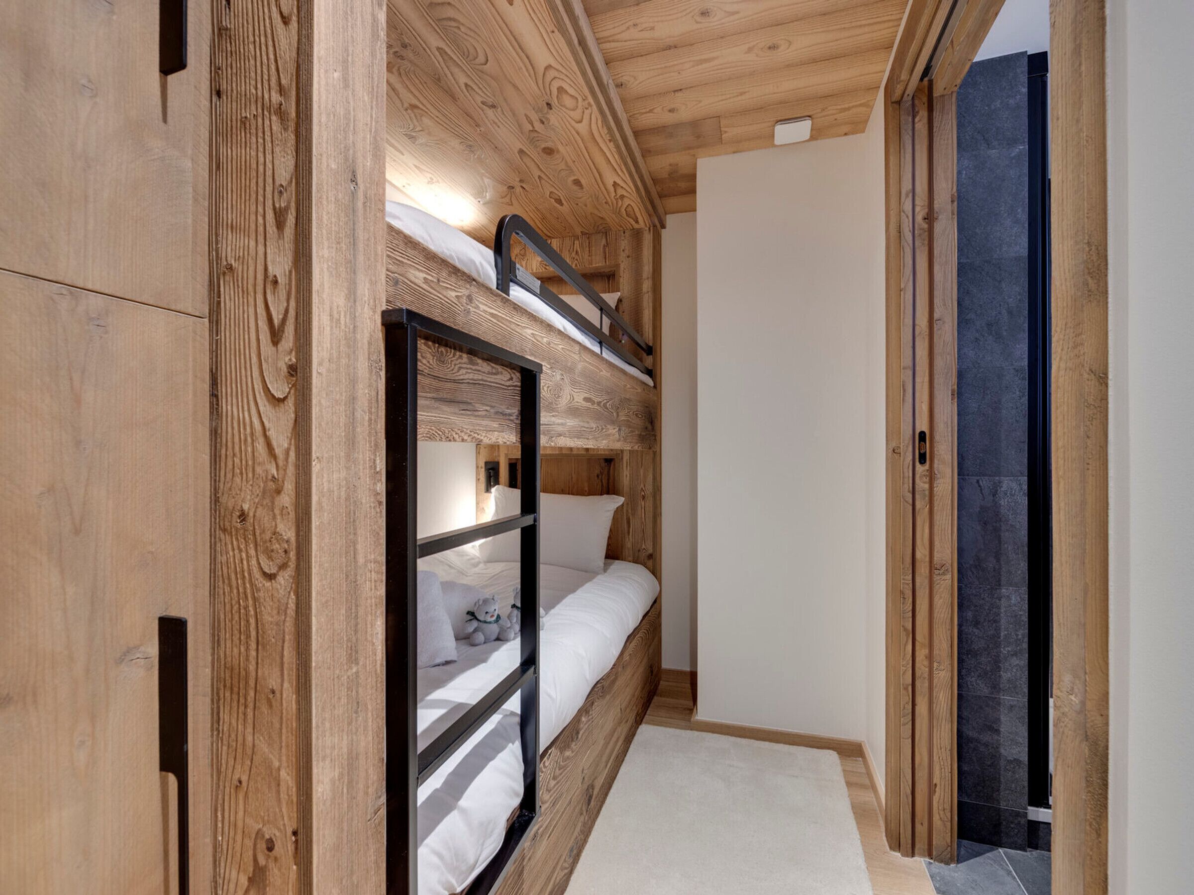Ein Stockbett mit Holz-Kopfteil, einer weißen Wand und einer Tür zu einem anderen Raum.