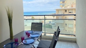 A balcony with a dining table set for two, overlooking a beach and buildings. - Charming T2 by the sea for 4 pers, terrace with view (Canet-Plage)