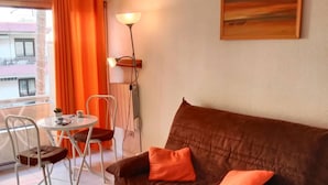 A cozy living area with a brown sofa, two orange cushions, a small round table, and two chairs. A floor lamp and a painting are on the wall. - Studio for 4 near the sea in Canet-en-Roussillon (Canet-en-Roussillon)