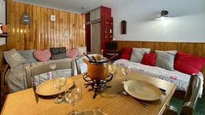 A cozy room with a wooden wall, a bed with red and grey pillows, a dining table set with plates, glasses, and a pot on a stove. - Ski-In Studio for 4 pers. with Parking (Le Dévoluy)