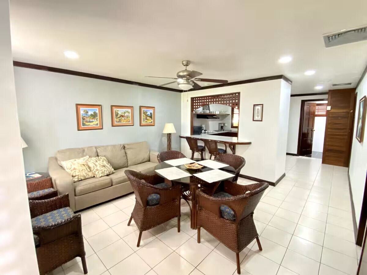 A living room with a sofa, dining table, and chairs, a kitchen area, and a ceiling fan.