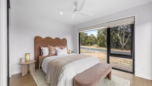 A bedroom with a large bed, a ceiling fan, a bedside table with a lamp, and a view of the outdoors.